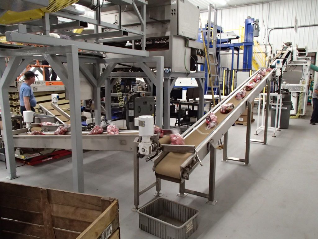 Food-packing hall — inclined belt conveyors carrying packaged meat products, stainless equipment and operators (third-party reference photo)
