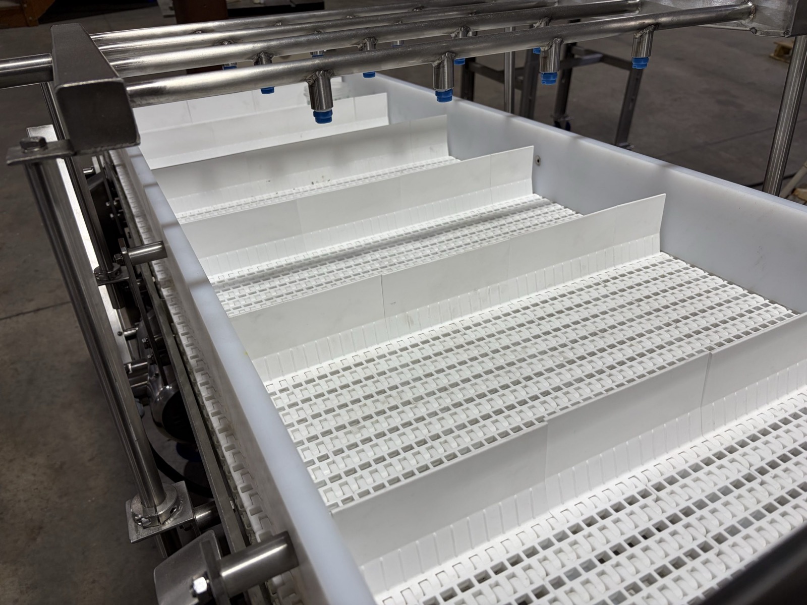 Plastic modular belt conveyor in a workshop — white modular belt in a stainless trough under a washdown spray header (third-party reference photo)