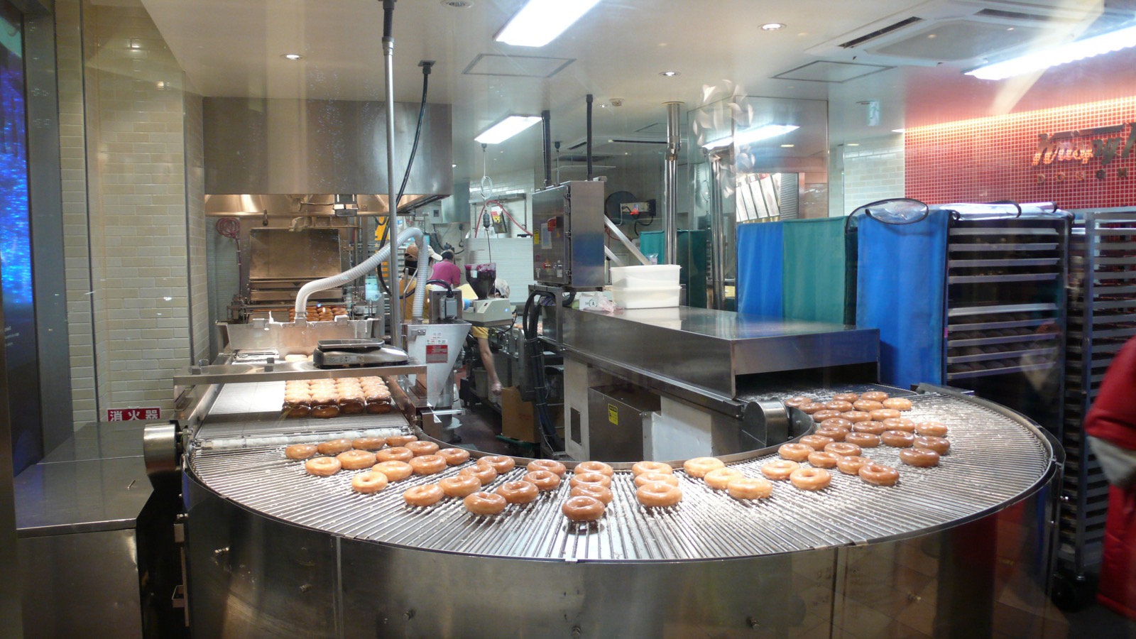 Bakery line — fried donuts carried on a metal mesh belt conveyor around a curved section, stainless steel framing (third-party reference photo)