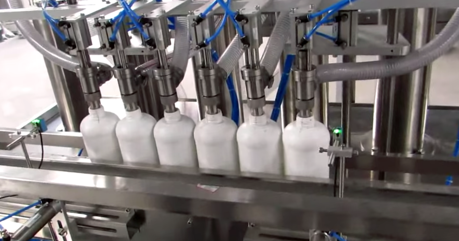 Daily-necessities filling line — six white plastic bottles lined up under the filling heads of a stainless filling machine (third-party reference photo)