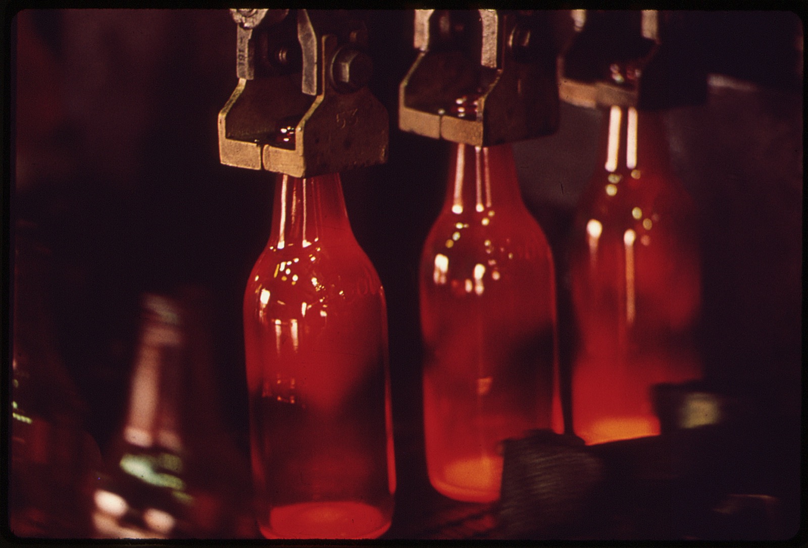 Glass production — newly moulded, still glowing glass bottles held by neck grippers on the line after forming (third-party reference photo)