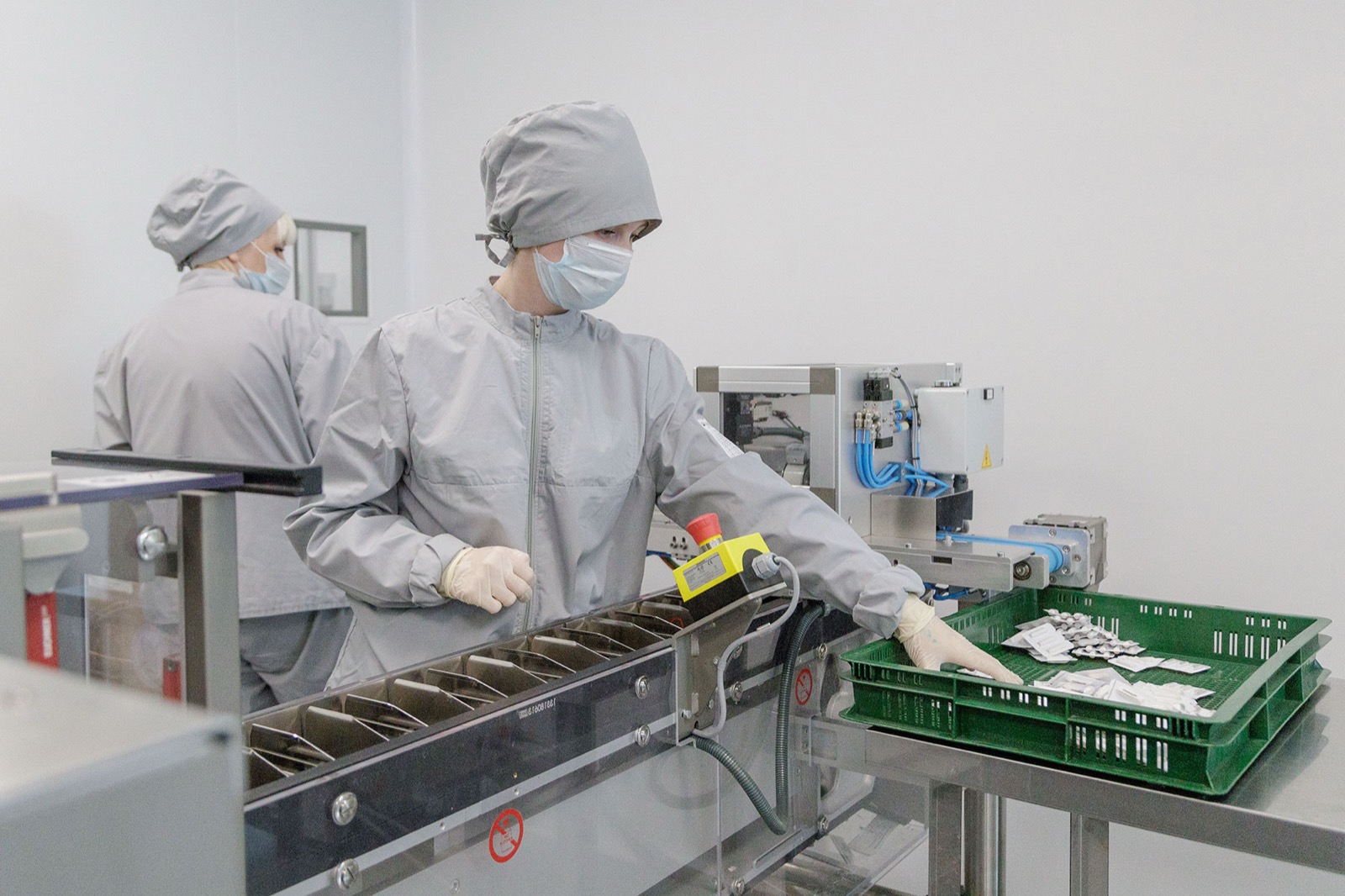 Pharmaceutical cleanroom — operators in cleanroom attire beside a divided conveyor carrying blister-packed medical products (third-party reference photo)