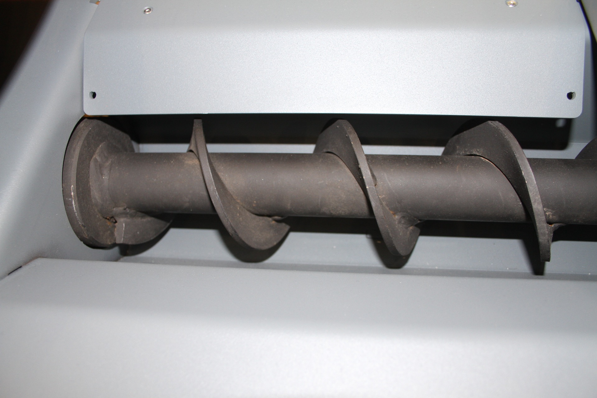 Third-party reference photo — close-up of a screw conveyor auger (helical flight on a shaft) inside a light metal trough; a wood-chip screw
